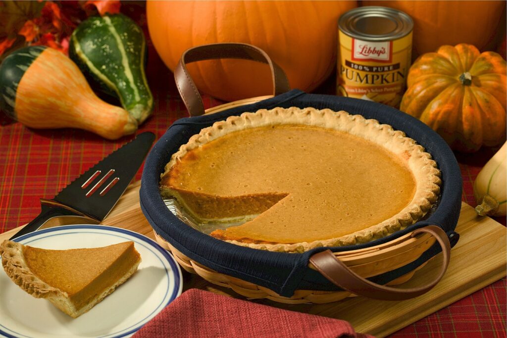 Pumpkin Pie with a piece missing and a piece of pumpkin pie sitting on a plate next to it. surrounding the pumpkin pie are thanksgiving meal items horns of plenty, pumpkins on a red tablecloth