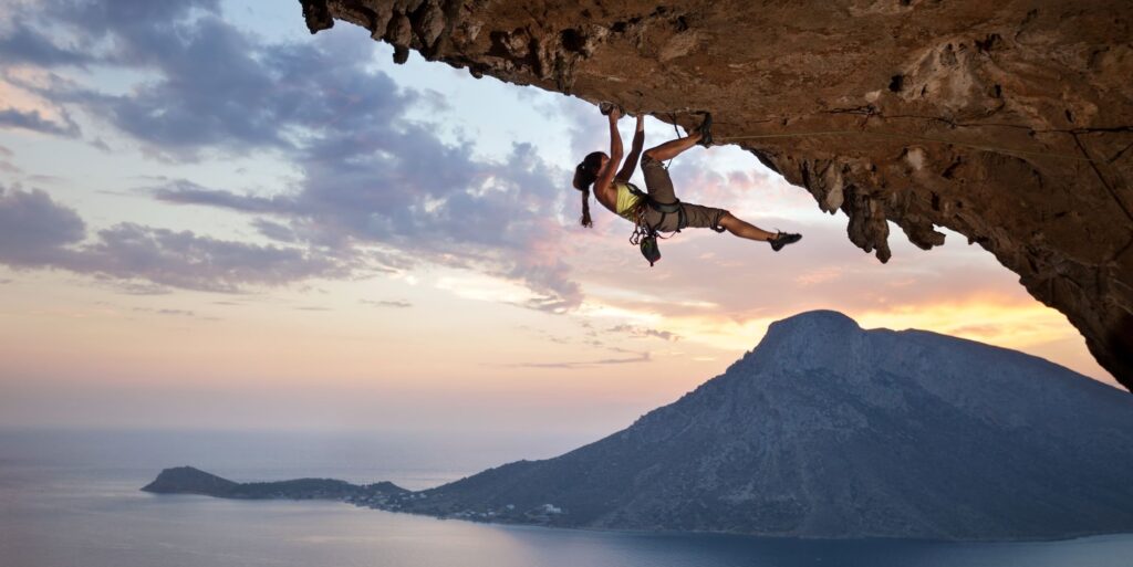 Female climber suspended horizontal on the underside of a mountain ledge. Beautiful mountain range and ocean in the background.Analogy shows business risk and opportunity